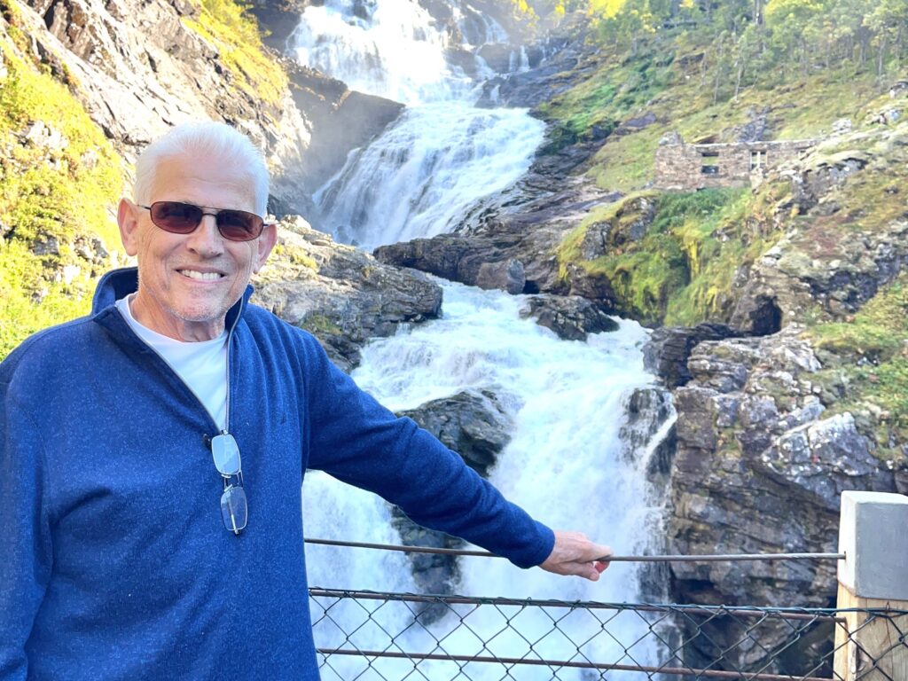 Rachel Friem's father standing in front of a waterfall during his 2022 trip to Norway.