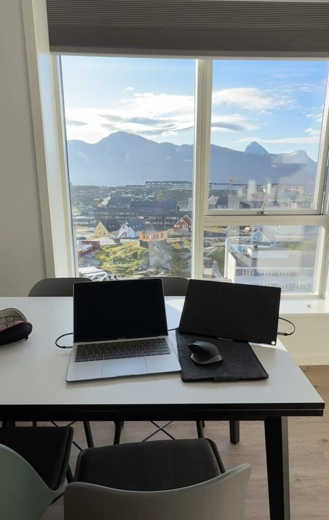 View from Hotel Aurora Apartments in Nuuk, Greenland, with a work table, large windows, colorful houses, and mountains in the distance.