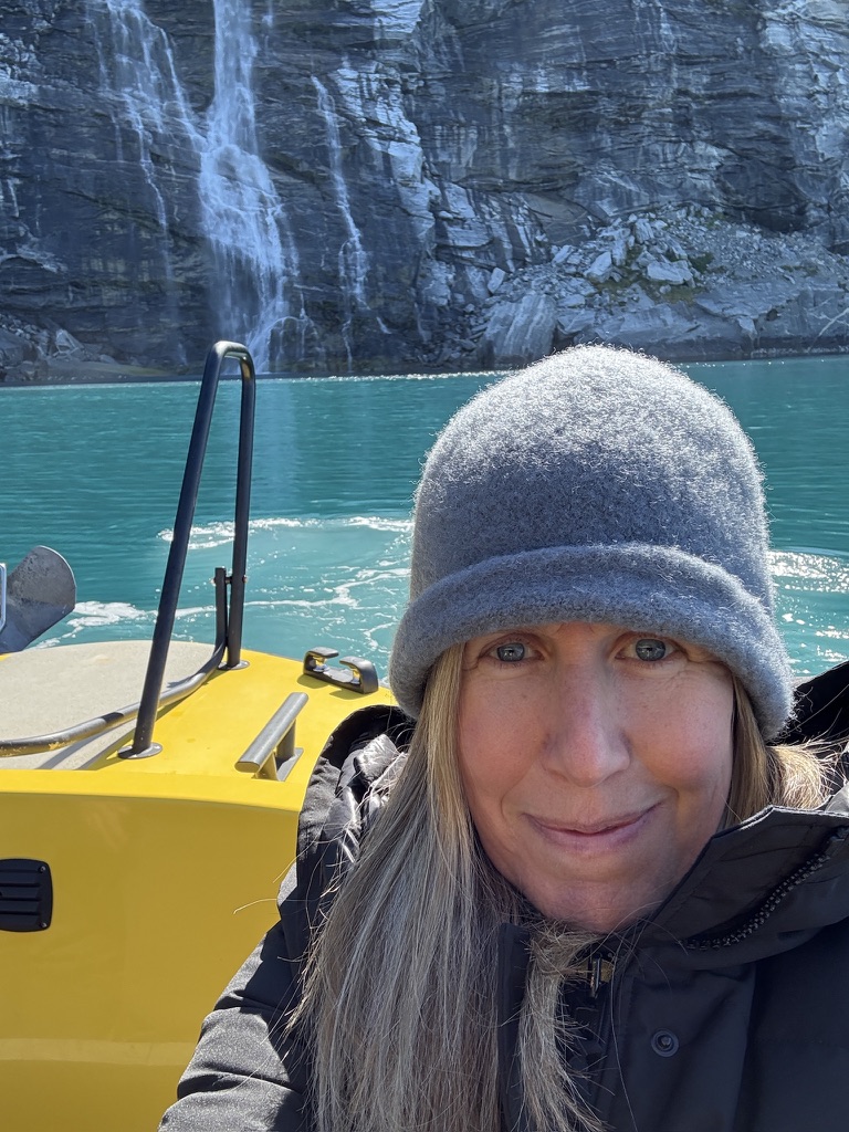 Rachel Friem bundled up on a Nuuk Water Taxi boat at the end of an Icefjord ride in Greenland with turquoise water and a rocky cliff behind her.