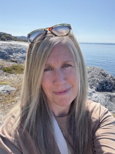 Rachel Friem taking a selfie on the rocky shoreline in Greenland with calm blue water behind her.